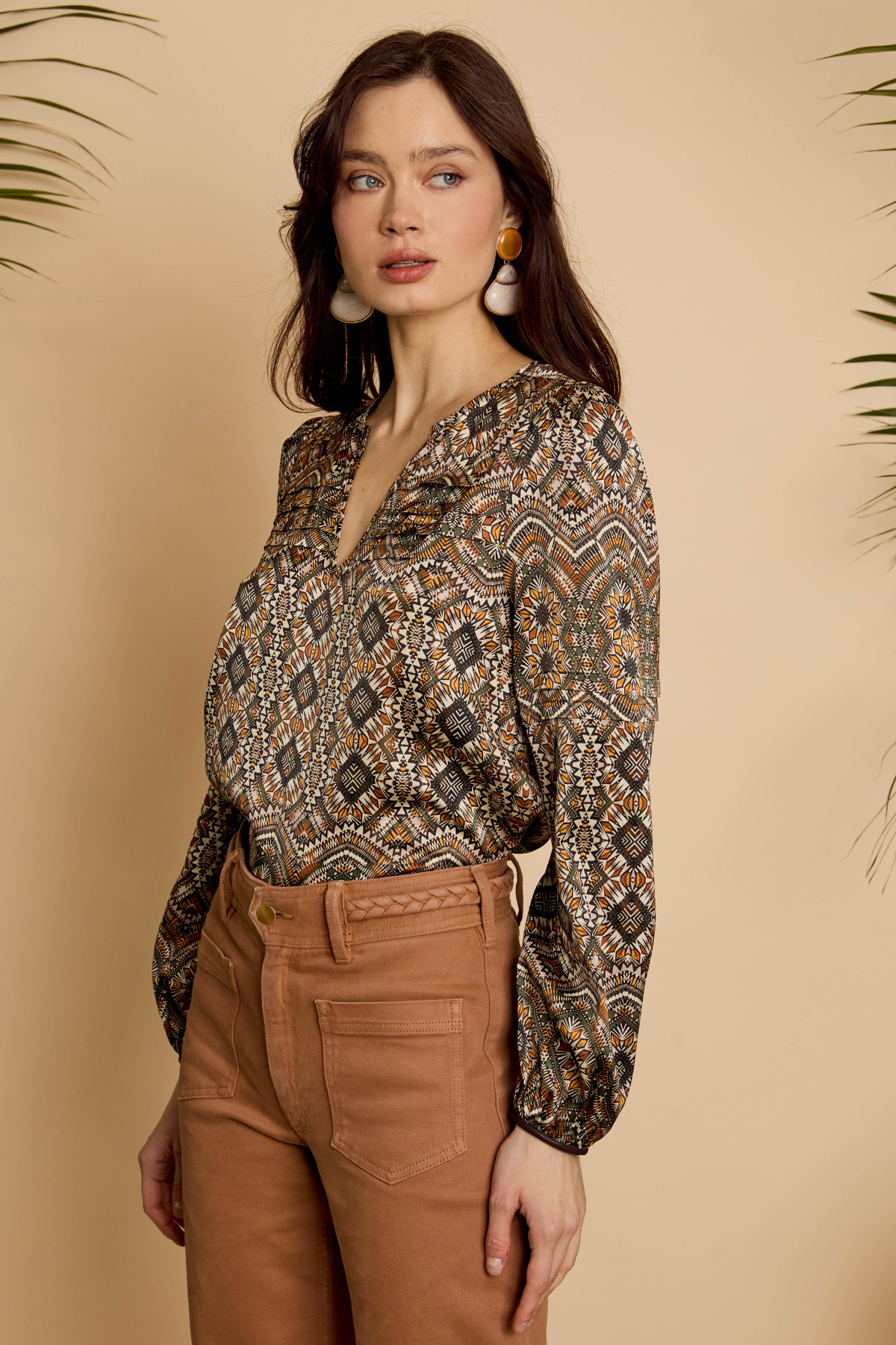 A woman modeling a long-sleeve V-neck blouse with a brown, beige, and black geometric pattern, paired with brown pants, set against a warm beige background with decorative palm leaves.