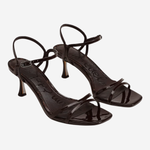 A pair of glossy black strappy high-heeled sandals with square toes and thin ankle straps, shown on a light gray background.