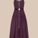A sleeveless midi dress in deep plum purple with a thin black belt at the waist featuring a gold buckle, and a central gold zipper running down the front. The dress has thin shoulder straps and a tiered skirt, shown against a solid beige background.