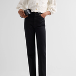 A model wearing a cream-colored double-breasted jacket with intricate eyelet lace sleeves and buttons, paired with black trousers and cream pointed-toe heels, holding a black clutch against a light gray studio background.