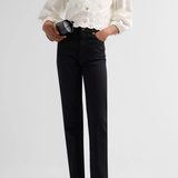 A model wearing a cream-colored double-breasted jacket with intricate eyelet lace sleeves and buttons, paired with black trousers and cream pointed-toe heels, holding a black clutch against a light gray studio background.