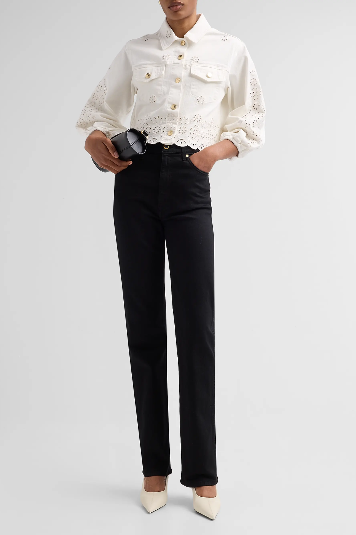 A model wearing a cream-colored double-breasted jacket with intricate eyelet lace sleeves and buttons, paired with black trousers and cream pointed-toe heels, holding a black clutch against a light gray studio background.