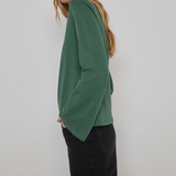 A woman wearing a sage green long-sleeve knit top and black pants, shown from a side-back angle against a plain light gray studio background.