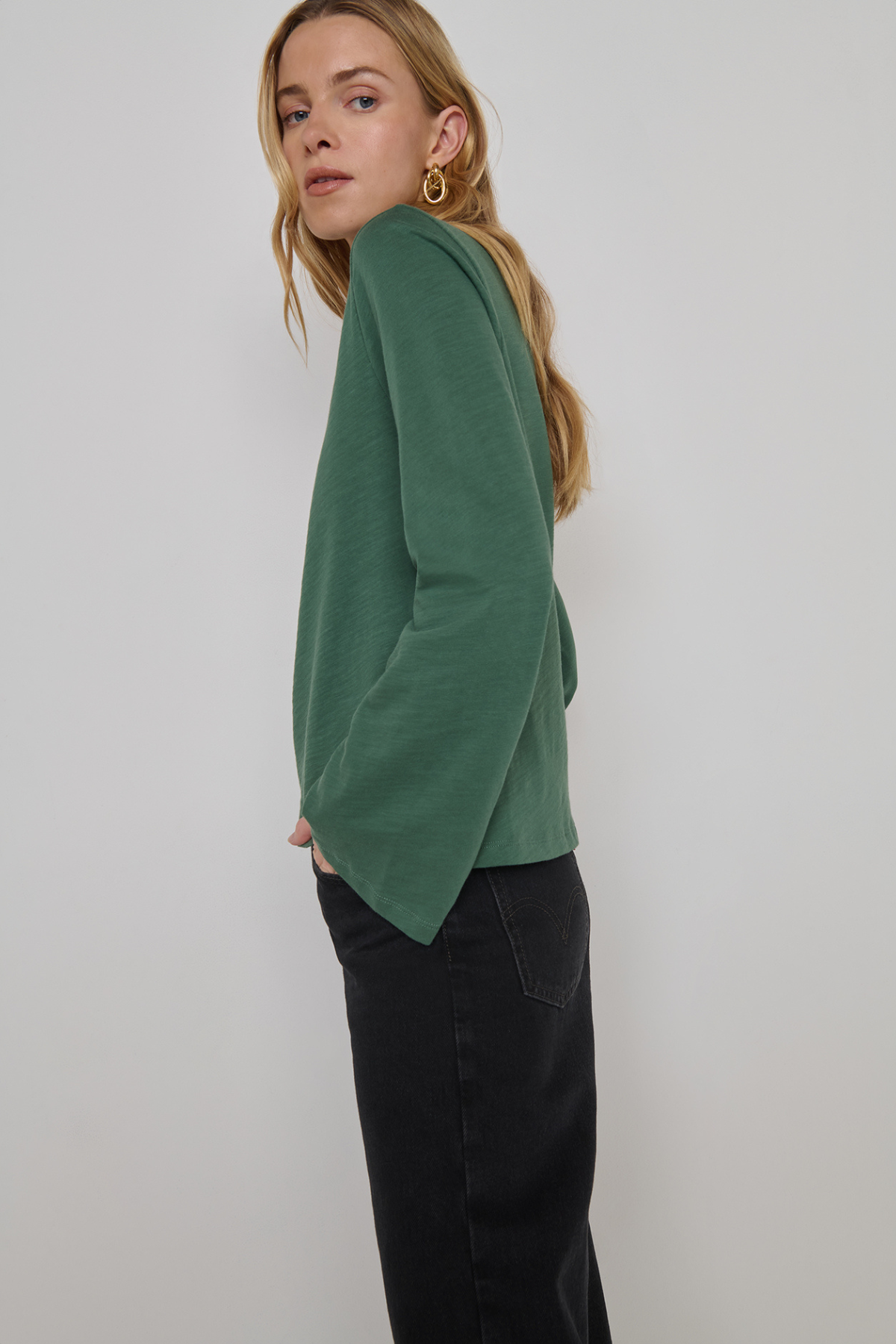 A woman wearing a sage green long-sleeve knit top and black pants, shown from a side-back angle against a plain light gray studio background.