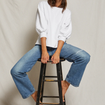 Woman sitting on a stool wearing a white blouse and blue jeans against a plain background