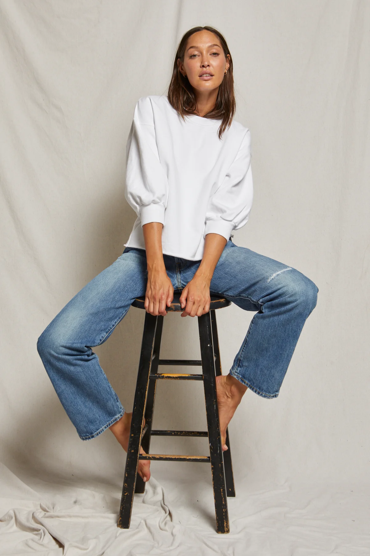 Woman sitting on a stool wearing a white blouse and blue jeans against a plain background