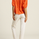 back of woman in orange eyelet top and white denim on tan background 