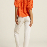 back of woman in orange eyelet top and white denim on tan background 