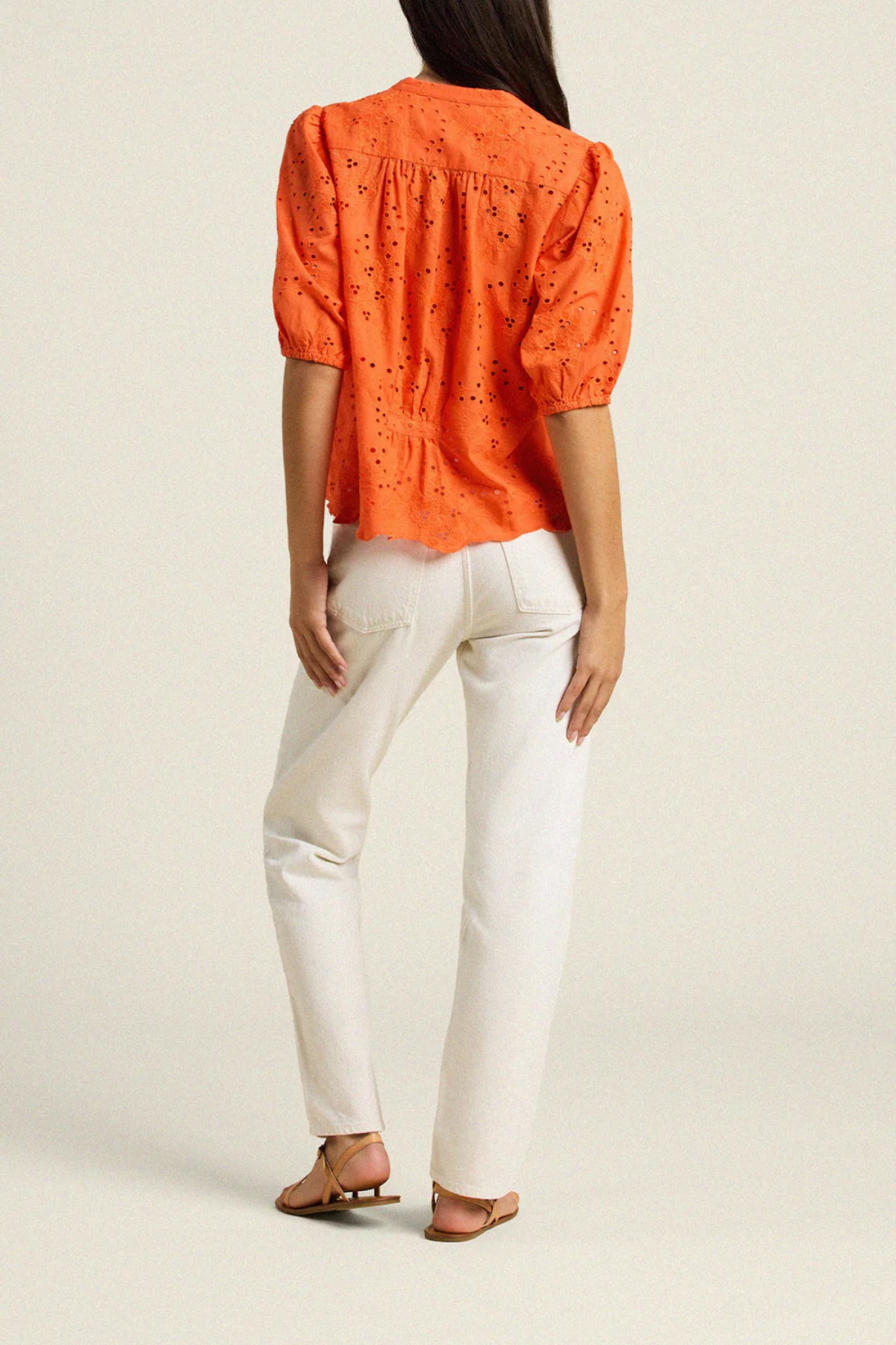 back of woman in orange eyelet top and white denim on tan background 