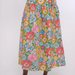 A multicolor floral midi skirt with a vibrant pink, yellow, blue, and green pattern, worn by a model with pink woven shoes against a plain white background.