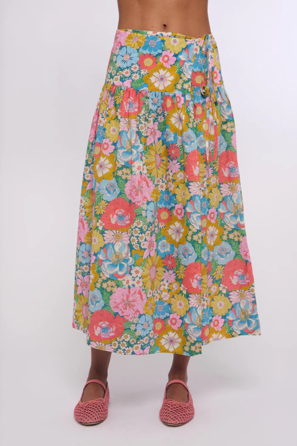 A multicolor floral midi skirt with a vibrant pink, yellow, blue, and green pattern, worn by a model with pink woven shoes against a plain white background.