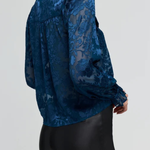 A woman wearing a dark blue sheer lace blouse with a high collar and long sleeves, paired with black pants, shown from a side-back angle against a light gray studio background.