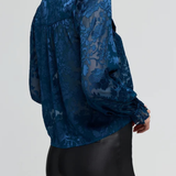 A woman wearing a dark blue sheer lace blouse with a high collar and long sleeves, paired with black pants, shown from a side-back angle against a light gray studio background.