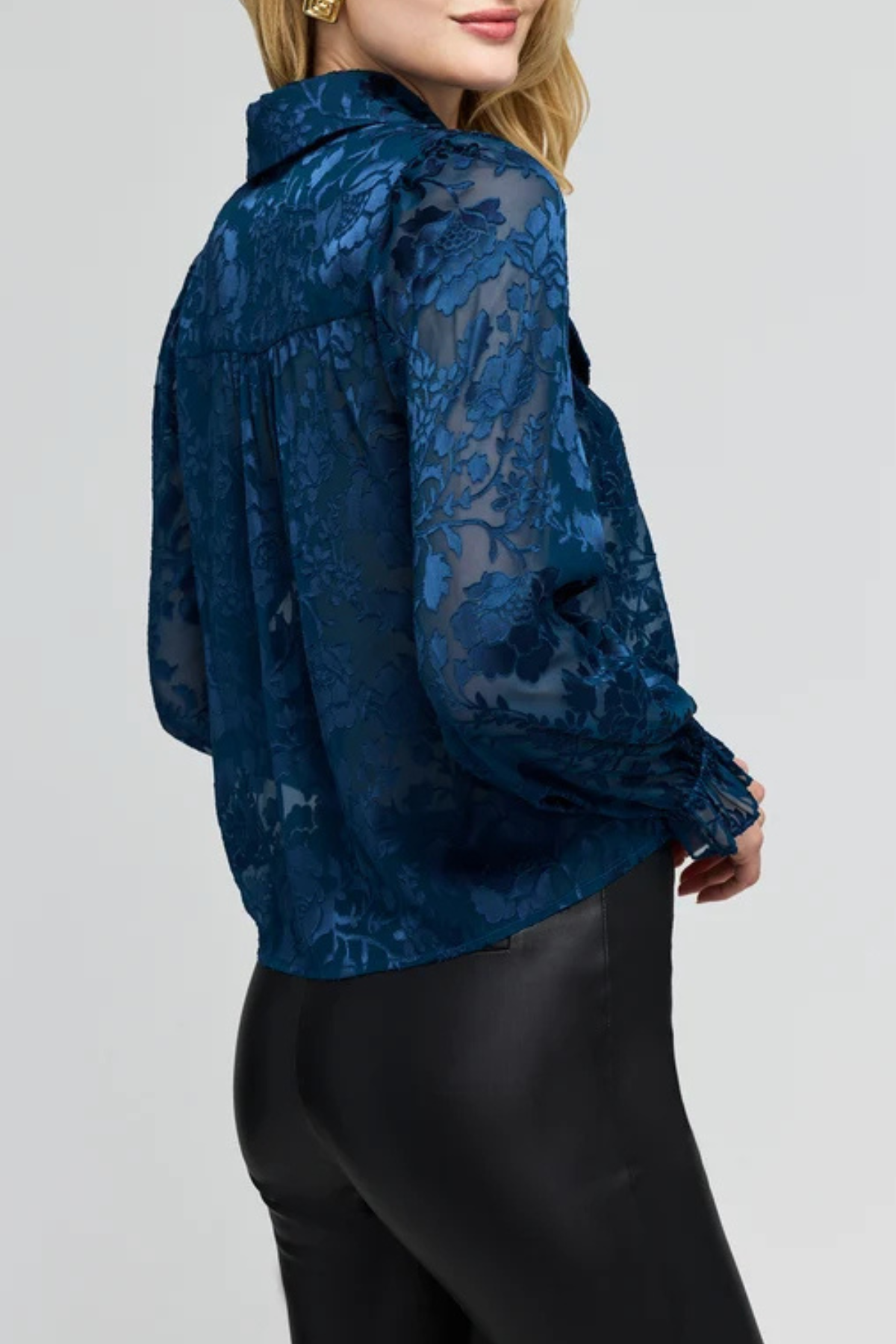 A woman wearing a dark blue sheer lace blouse with a high collar and long sleeves, paired with black pants, shown from a side-back angle against a light gray studio background.