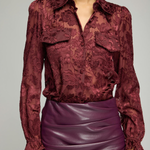 A woman wearing a long-sleeve burgundy lace blouse with a collar and chest pockets, paired with a ruched purple faux-leather skirt, against a plain light gray studio background.