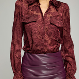 A woman wearing a long-sleeve burgundy lace blouse with a collar and chest pockets, paired with a ruched purple faux-leather skirt, against a plain light gray studio background.