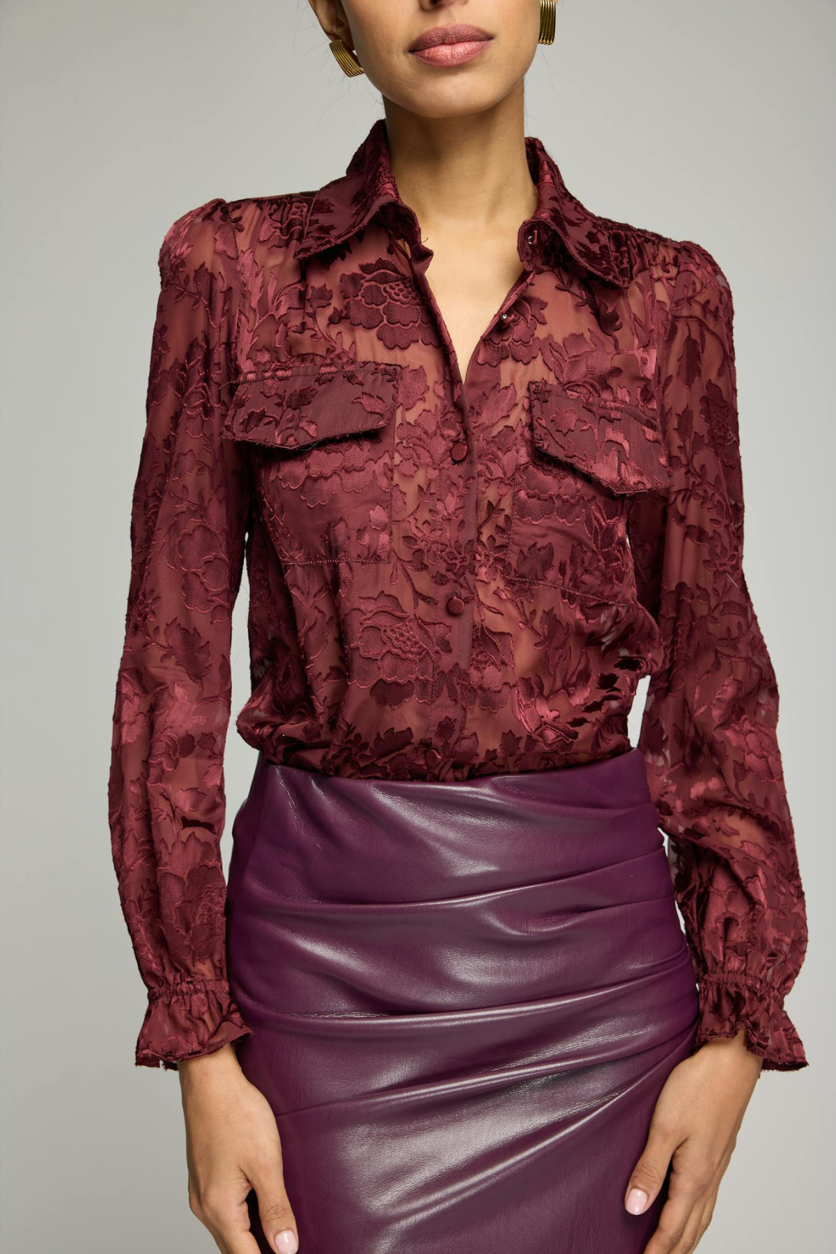 A woman wearing a long-sleeve burgundy lace blouse with a collar and chest pockets, paired with a ruched purple faux-leather skirt, against a plain light gray studio background.