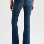 Rear view of a model wearing blue denim flare jeans with decorative embroidery on the back pockets, paired with a black top and black heels, against a light gray studio background.