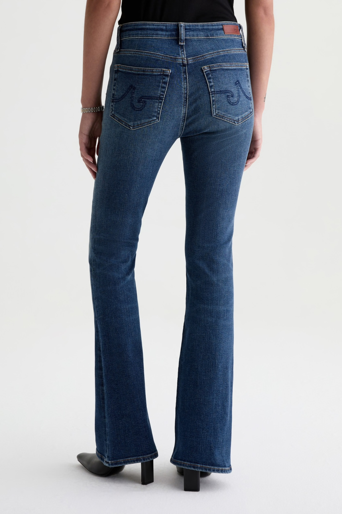 Rear view of a model wearing blue denim flare jeans with decorative embroidery on the back pockets, paired with a black top and black heels, against a light gray studio background.