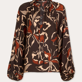 A long-sleeve blouse with a brown base and an abstract floral pattern in beige and rust orange, featuring a tie-neck detail and gathered cuffs, shown against a light gray background.
