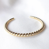 A gold twisted rope cuff bracelet resting on a soft, white textured fabric background.