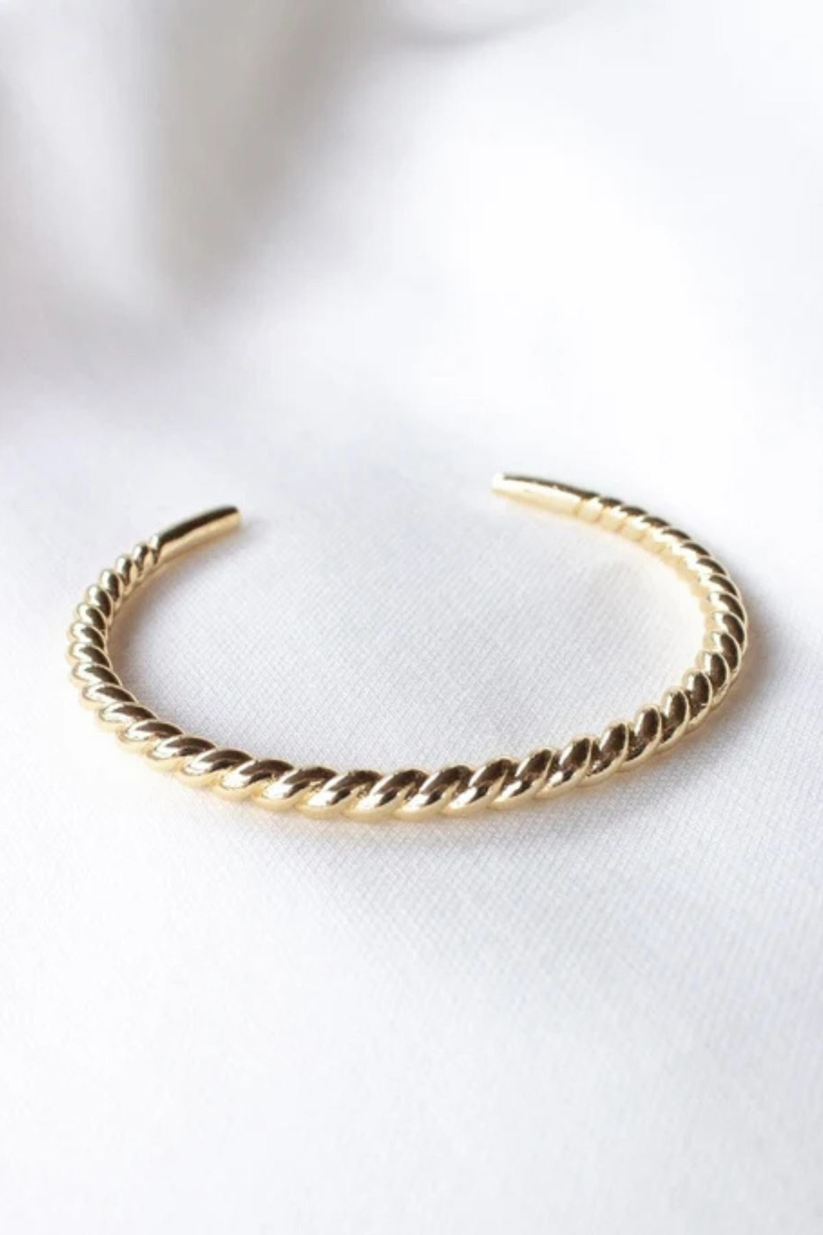 A gold twisted rope cuff bracelet resting on a soft, white textured fabric background.