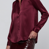 A woman wearing a long-sleeve satin button-up blouse in deep burgundy, paired with glossy dark burgundy pants, against a light gray studio background.