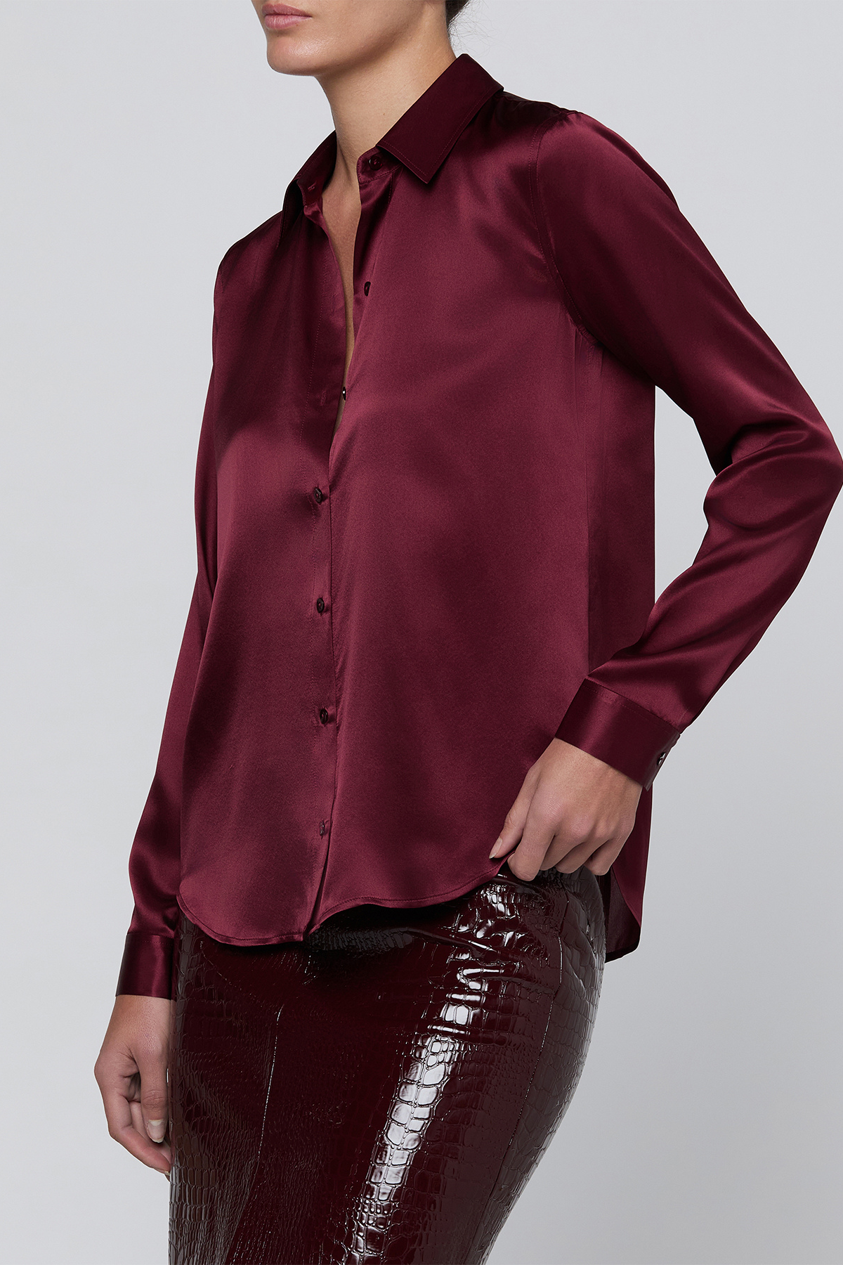 A woman wearing a long-sleeve satin button-up blouse in deep burgundy, paired with glossy dark burgundy pants, against a light gray studio background.