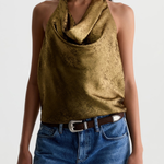 A woman wearing a metallic gold halter top with a textured, crinkled finish, paired with blue jeans and a brown belt, against a plain light gray background.