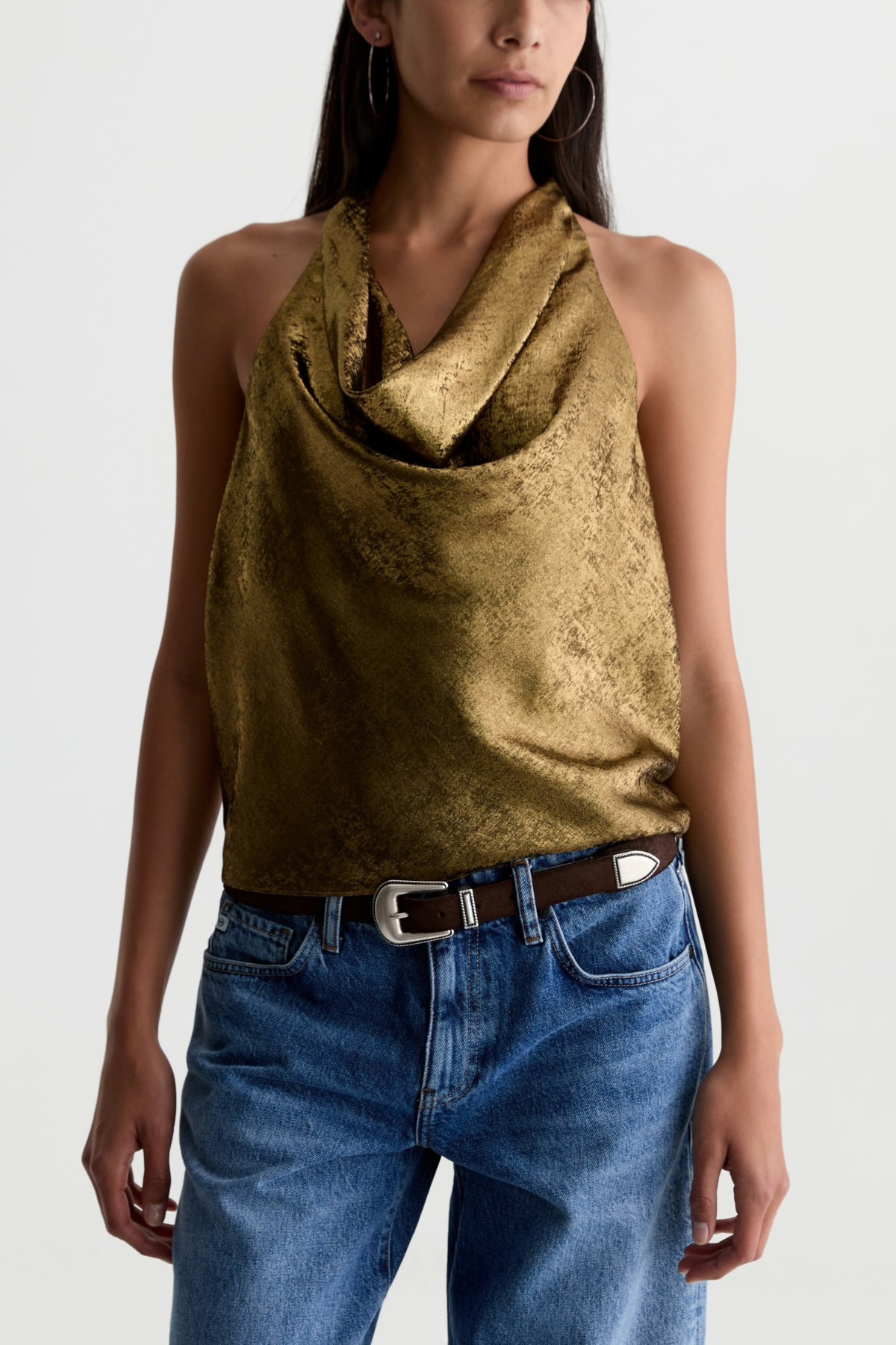 A woman wearing a metallic gold halter top with a textured, crinkled finish, paired with blue jeans and a brown belt, against a plain light gray background.