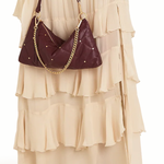 A person wearing a long cream ruffled dress and black pointed shoes carries a dark burgundy crescent-shaped shoulder bag with gold-tone chain and star details against a plain white background.