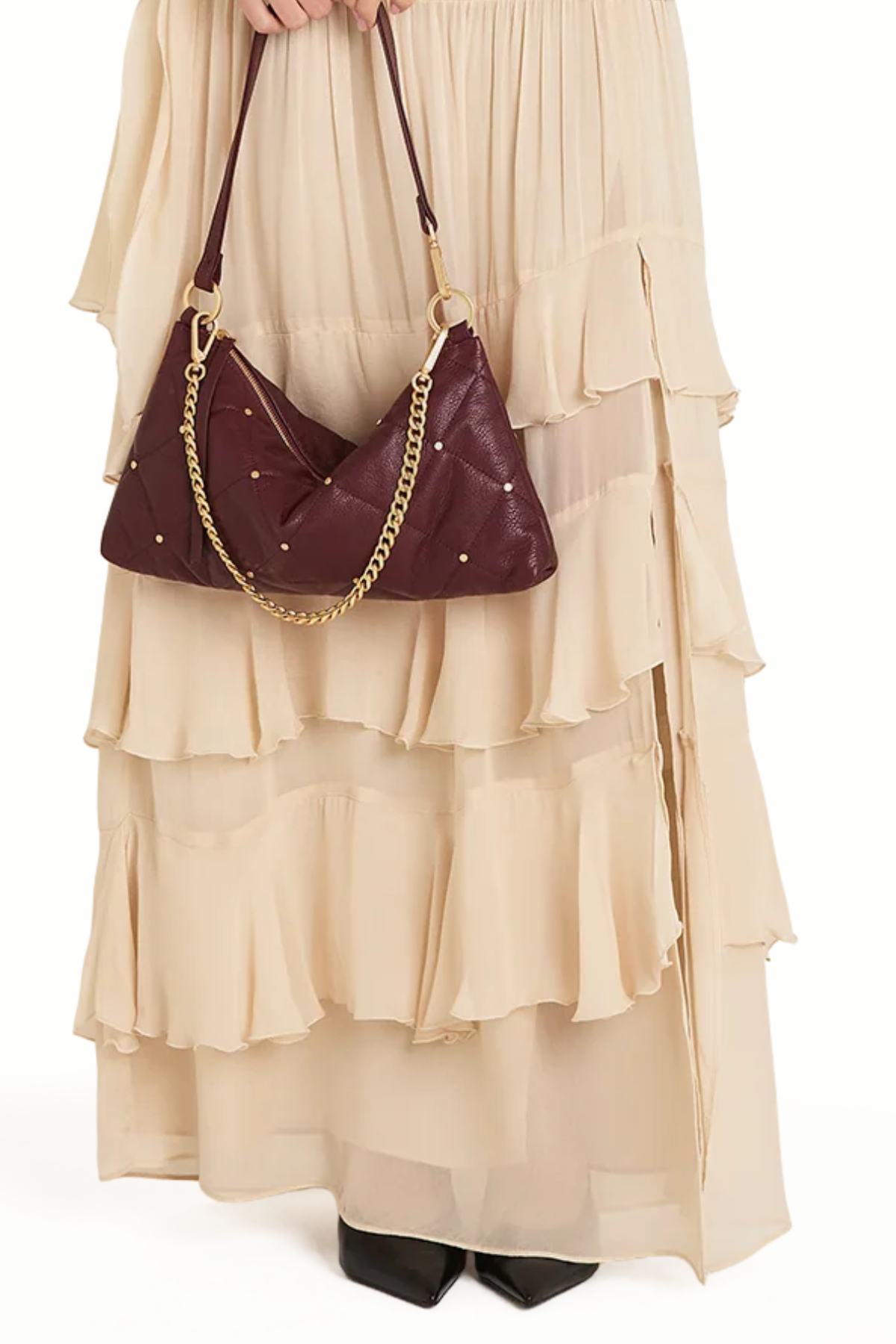 A person wearing a long cream ruffled dress and black pointed shoes carries a dark burgundy crescent-shaped shoulder bag with gold-tone chain and star details against a plain white background.