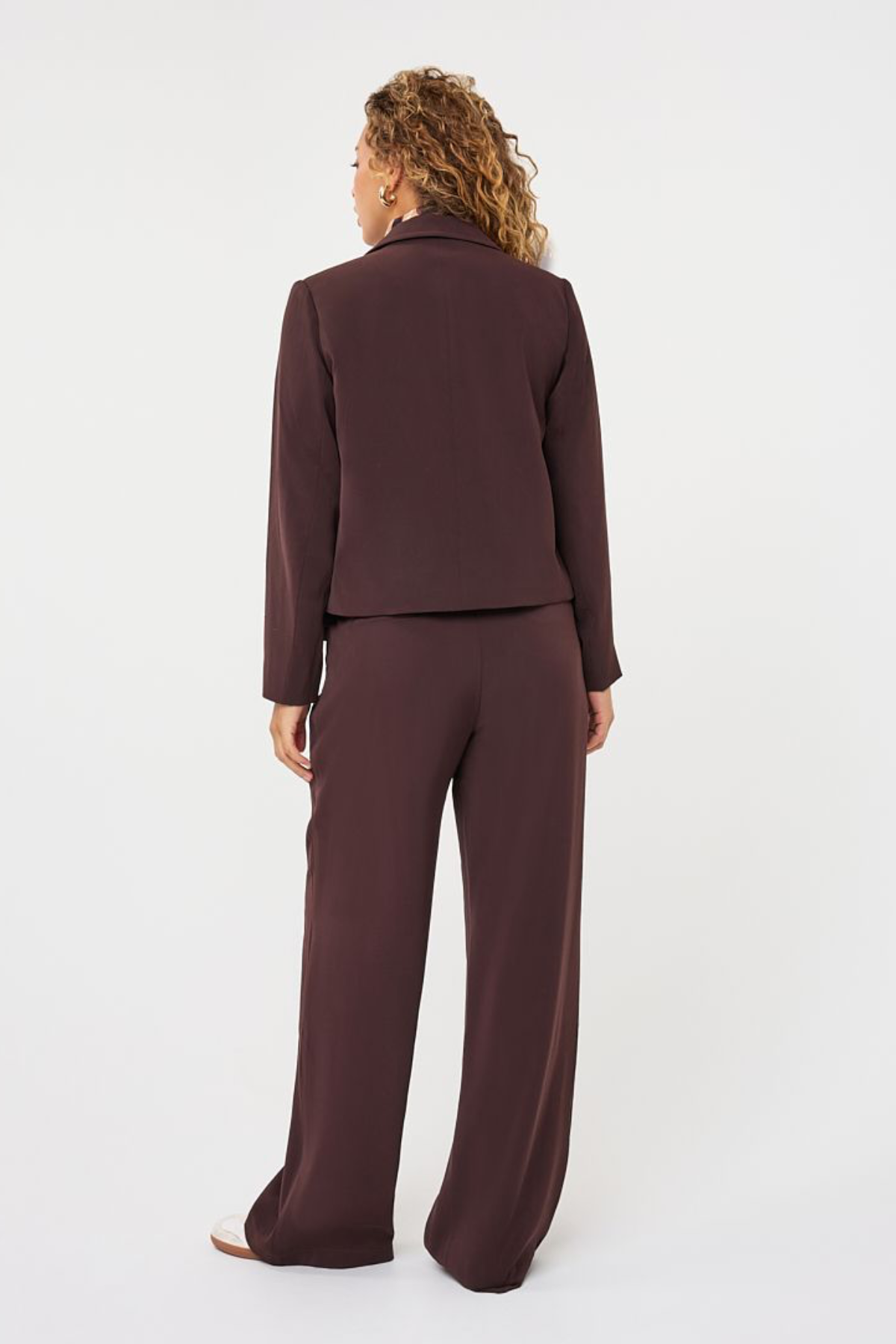 A woman shown from the back wearing a matching dark plum-brown two-piece suit, consisting of a cropped long-sleeve jacket and wide-leg trousers, against a plain white studio background.