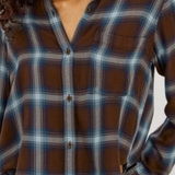 A close-up of a woman wearing a brown, blue, and gray plaid button-up shirt with dark denim jeans.