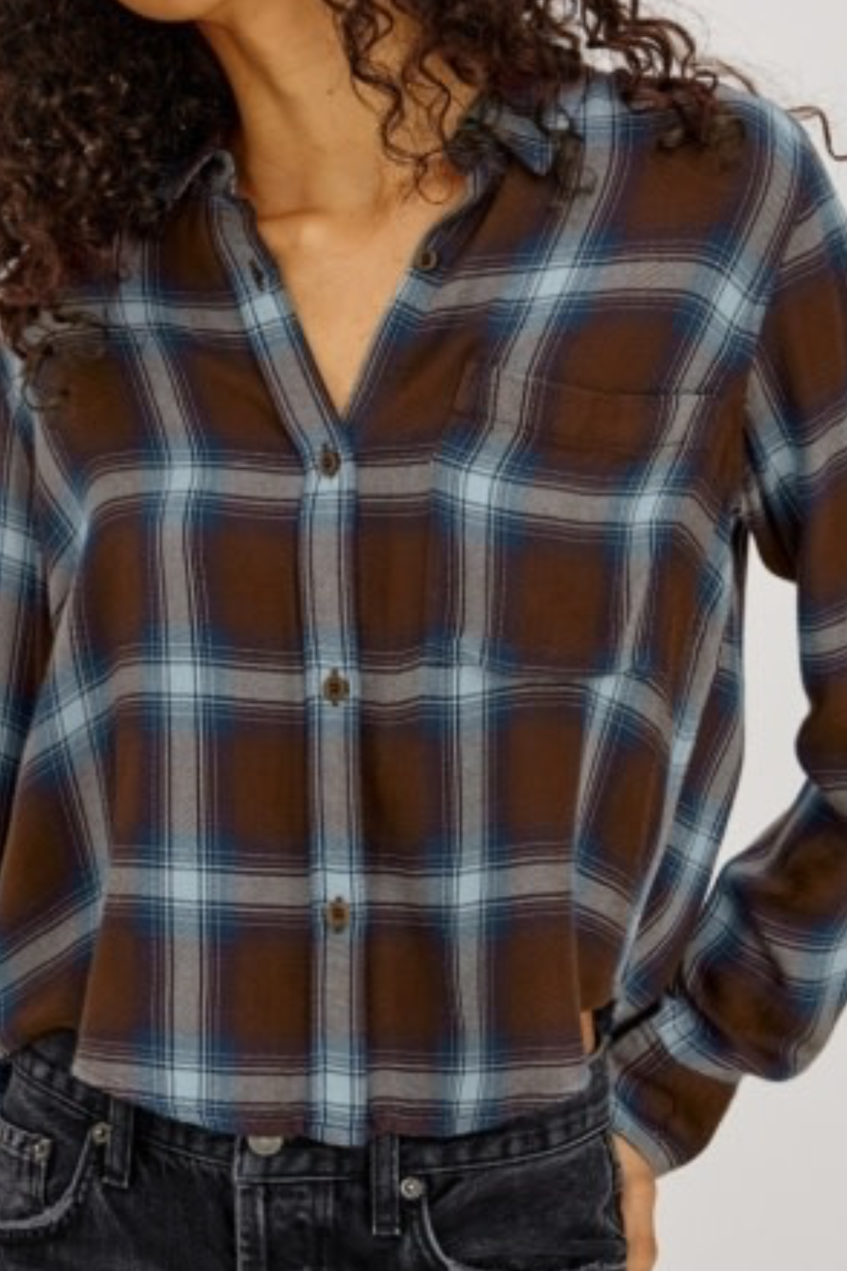 A close-up of a woman wearing a brown, blue, and gray plaid button-up shirt with dark denim jeans.