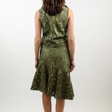 A woman viewed from behind wearing a sleeveless olive green dress with an intricate paisley pattern and a tiered ruffled skirt, paired with brown platform sandals, against a plain white studio background.