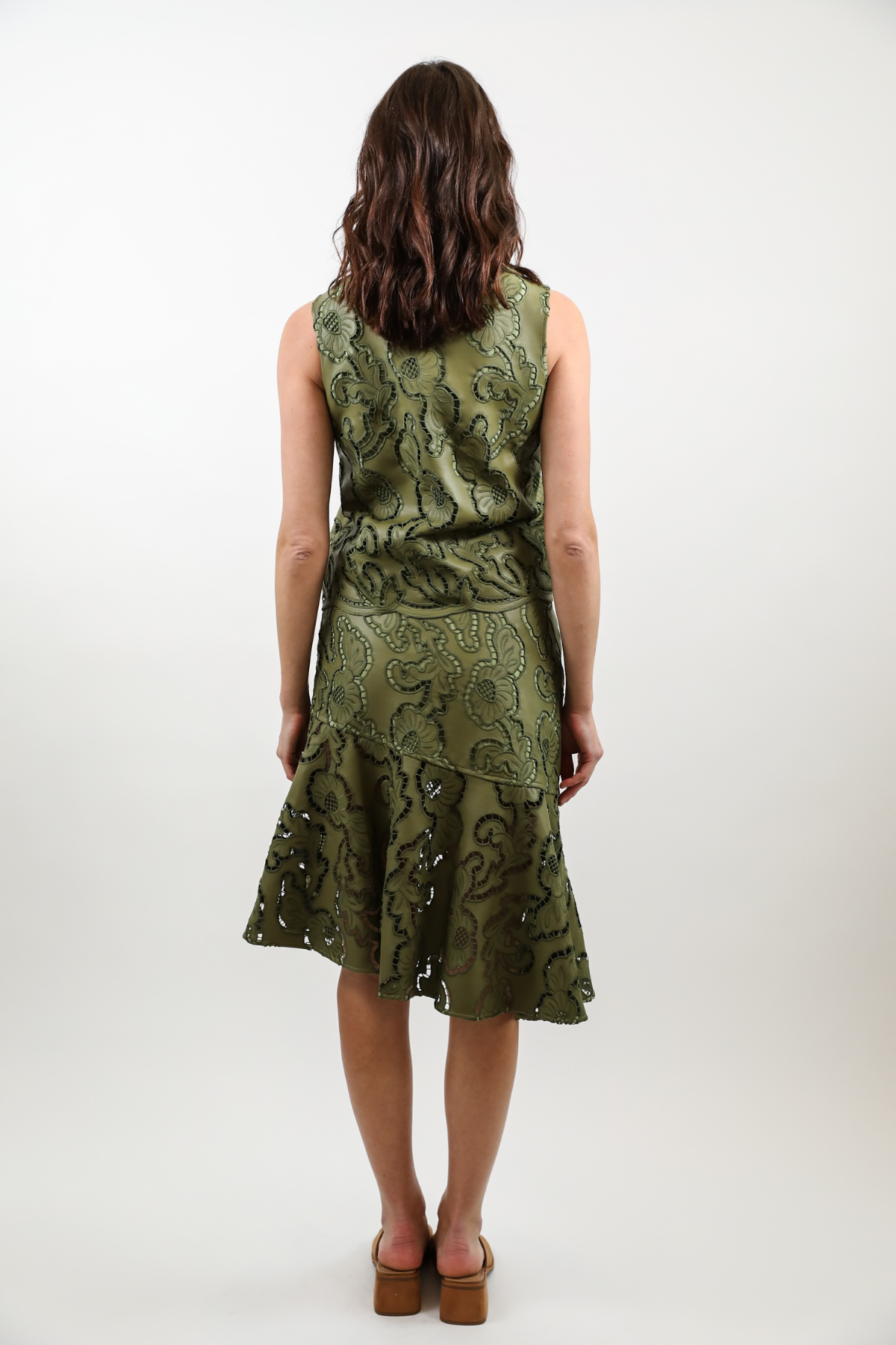 A woman viewed from behind wearing a sleeveless olive green dress with an intricate paisley pattern and a tiered ruffled skirt, paired with brown platform sandals, against a plain white studio background.
