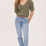 A woman modeling an olive green short-sleeve knit top with light-wash blue jeans and tan ankle boots, standing against a plain light beige studio background.