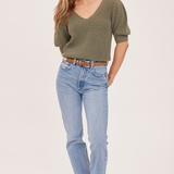 A woman modeling an olive green short-sleeve knit top with light-wash blue jeans and tan ankle boots, standing against a plain light beige studio background.