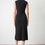 A woman viewed from the back wearing a sleeveless black pleated midi dress with a mock neck, paired with black pointed-toe heels, set against a plain light gray wall and concrete floor.