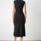 A woman viewed from the back wearing a sleeveless black pleated midi dress with a mock neck, paired with black pointed-toe heels, set against a plain light gray wall and concrete floor.