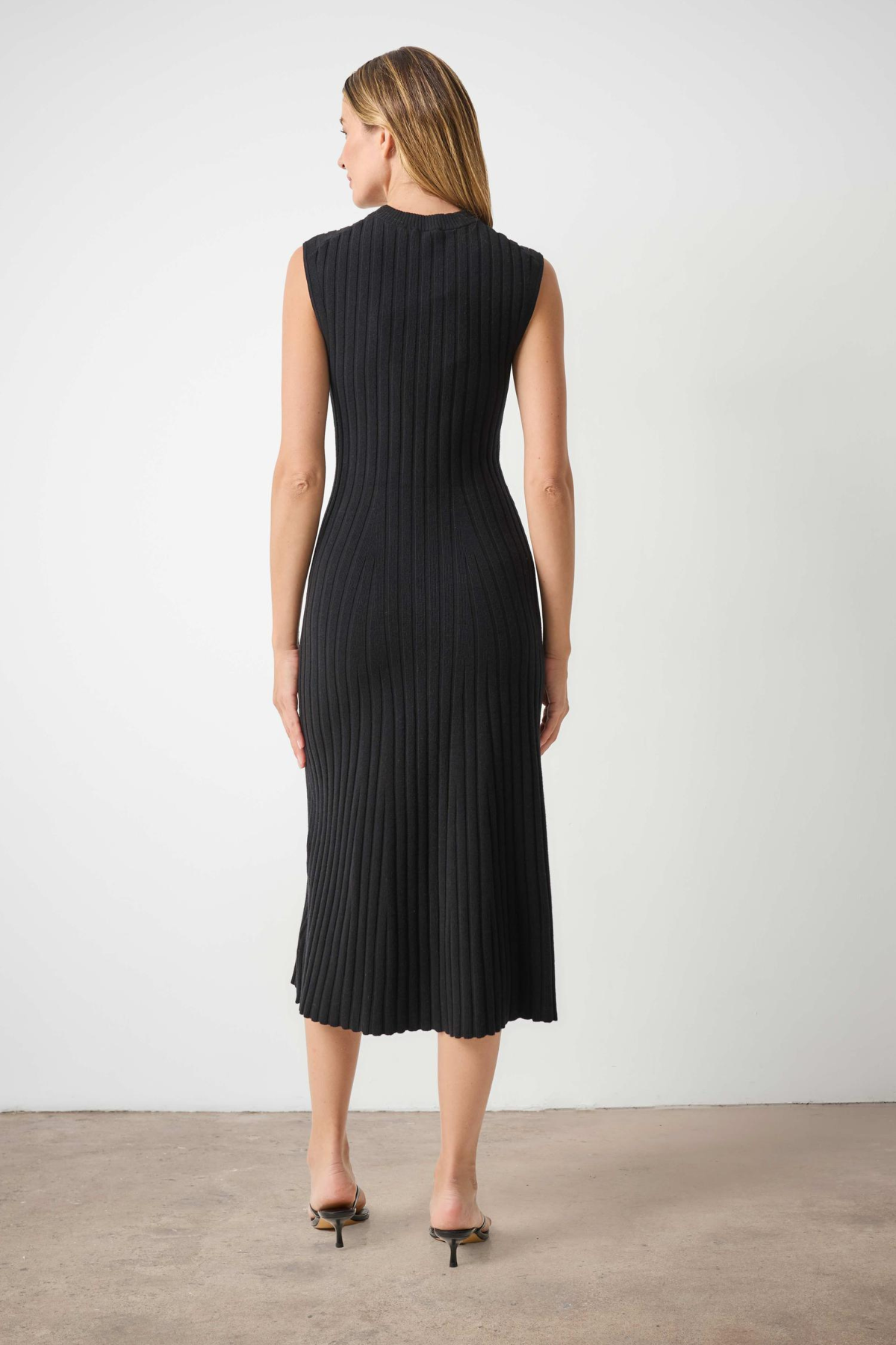A woman viewed from the back wearing a sleeveless black pleated midi dress with a mock neck, paired with black pointed-toe heels, set against a plain light gray wall and concrete floor.