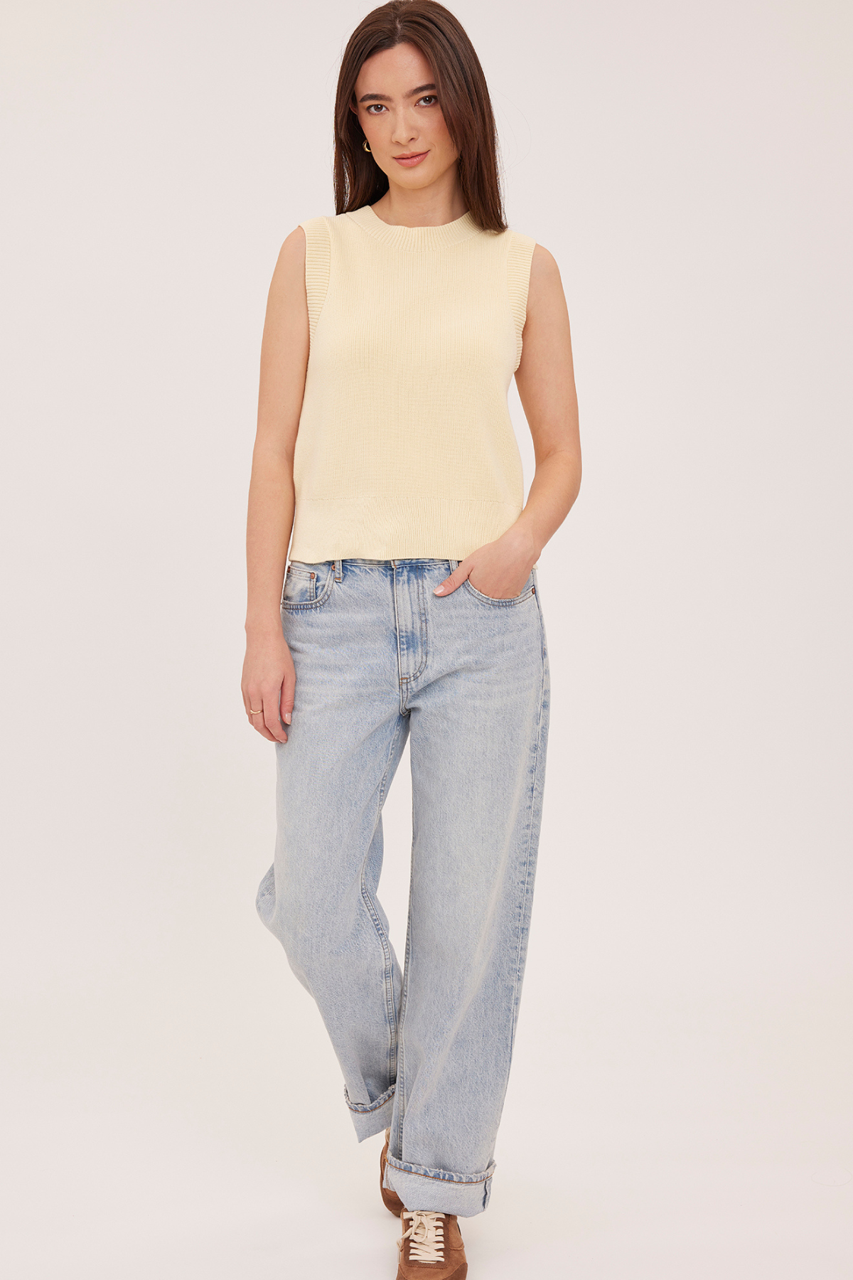 A woman modeling a pale yellow sleeveless knit top with light-wash wide-leg jeans and brown shoes, standing against a plain light background.