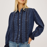 A woman wearing a dark blue denim button-up shirt with vertical embroidered stripes and light-wash blue jeans, photographed against a plain light gray studio background.
