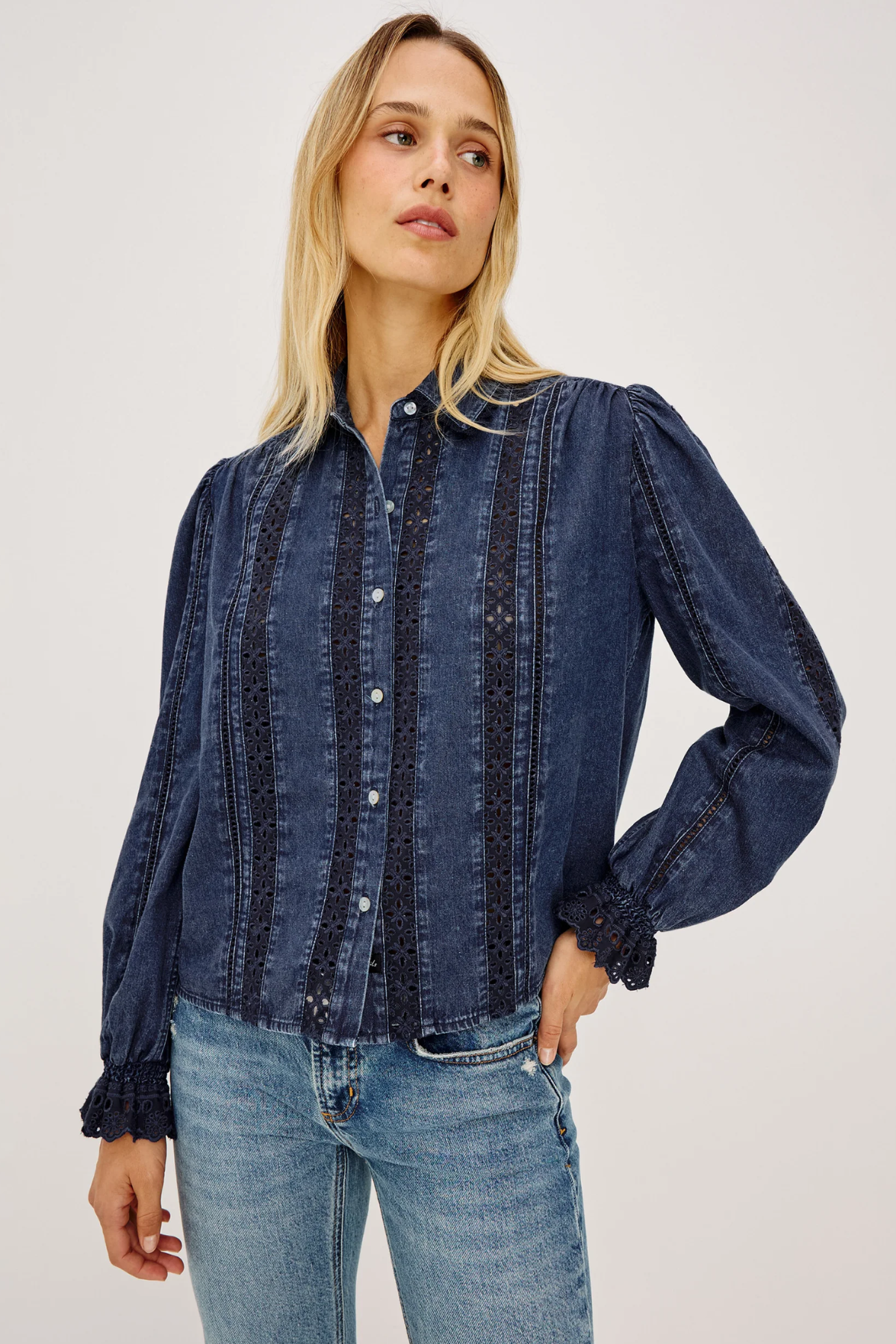 A woman wearing a dark blue denim button-up shirt with vertical embroidered stripes and light-wash blue jeans, photographed against a plain light gray studio background.