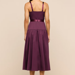 A woman viewed from the back wearing a sleeveless midi dress in deep plum purple with satin straps and a matching waistband, standing against a plain beige studio background.