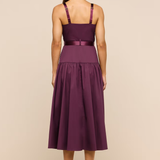 A woman viewed from the back wearing a sleeveless midi dress in deep plum purple with satin straps and a matching waistband, standing against a plain beige studio background.