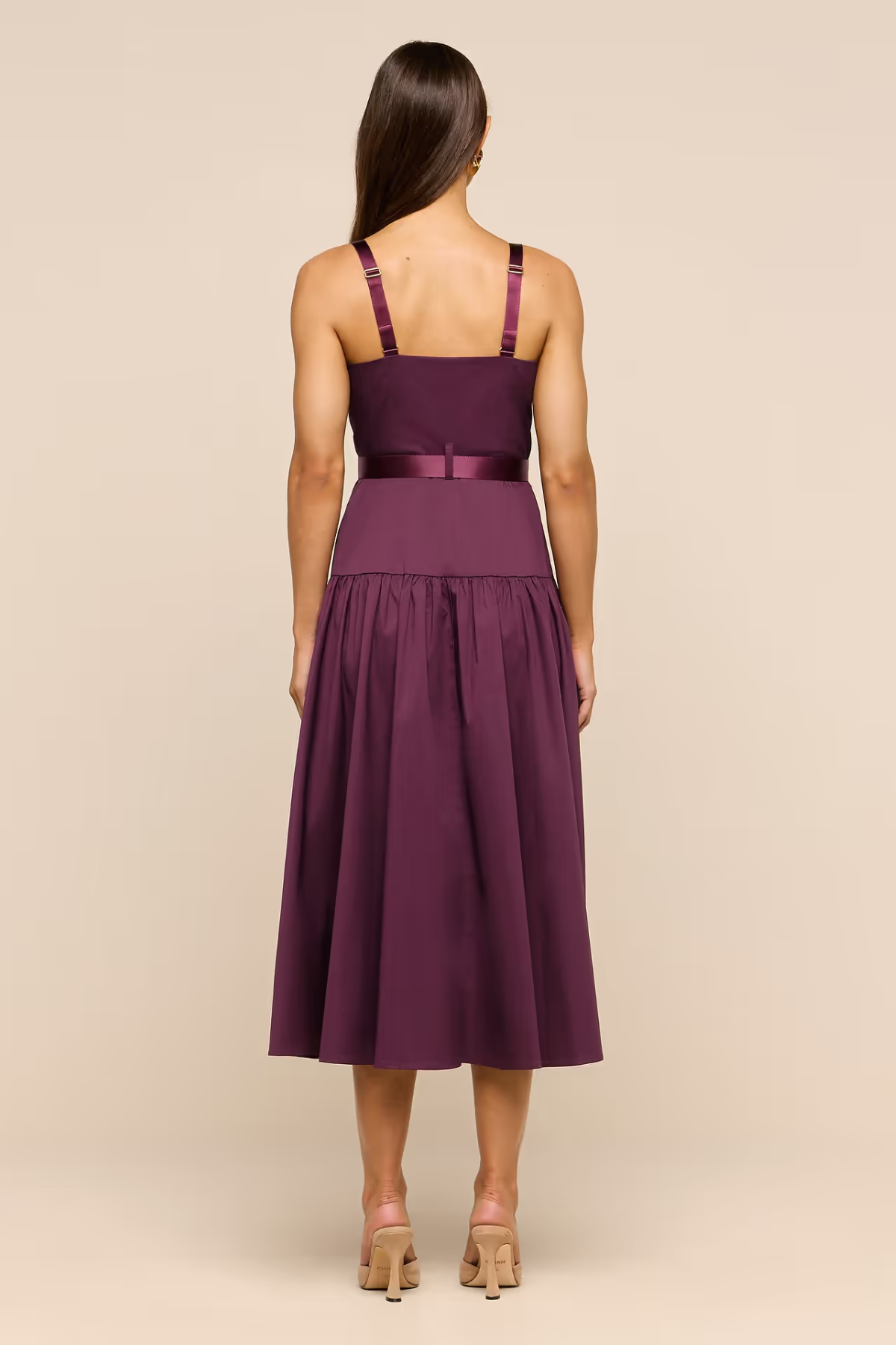 A woman viewed from the back wearing a sleeveless midi dress in deep plum purple with satin straps and a matching waistband, standing against a plain beige studio background.