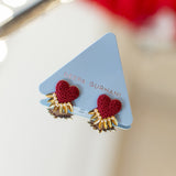 A pair of red beaded heart stud earrings with gold sunburst details, displayed on a light blue triangular card printed with the brand name 'DETTA SURNANI'.