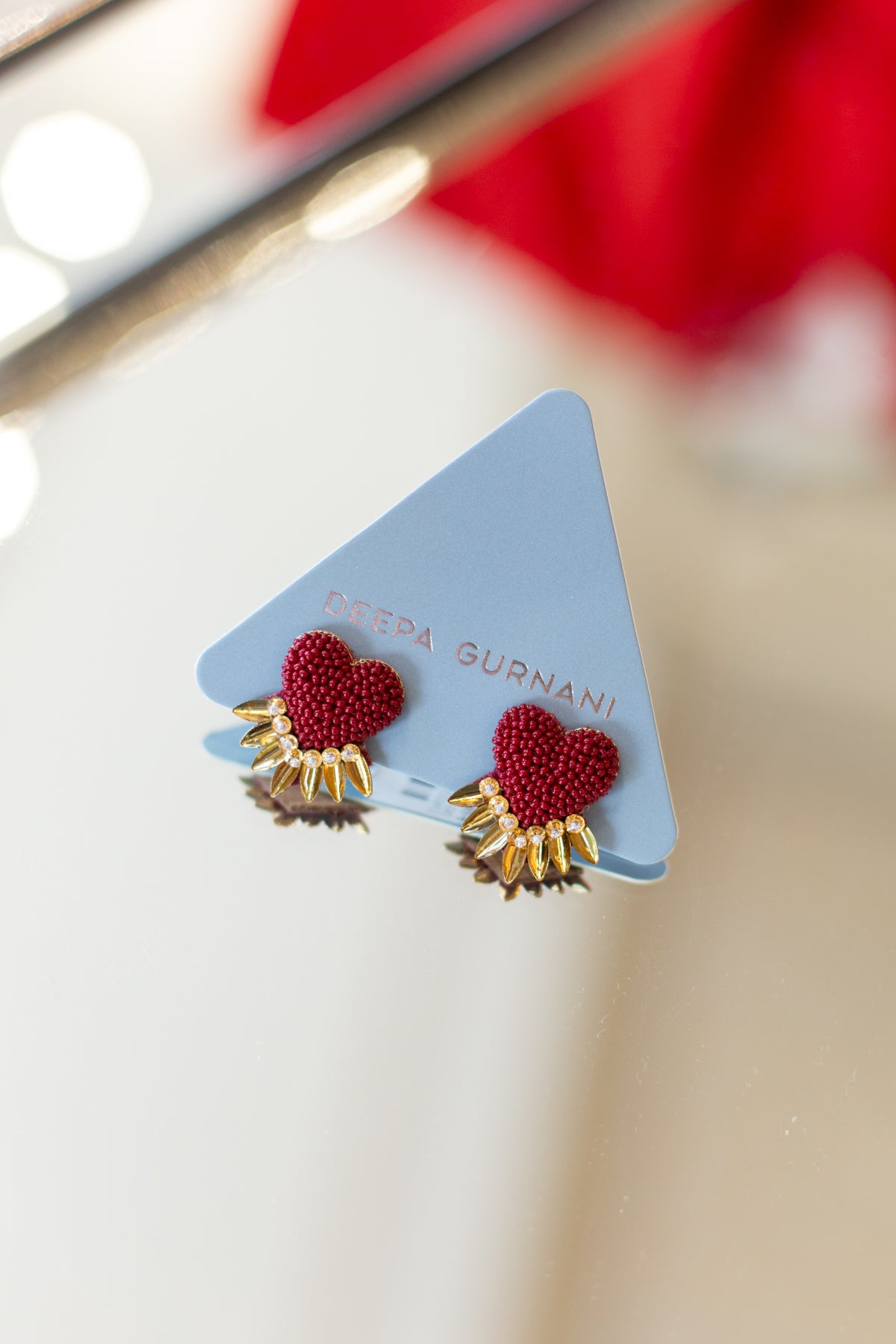 A pair of red beaded heart stud earrings with gold sunburst details, displayed on a light blue triangular card printed with the brand name 'DETTA SURNANI'.